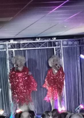 Zwei Drag-Künstlerinnen in roten Paillettenkleidern singen auf einer Bühne.